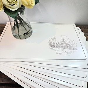 May include: A stack of white placemats with a black border and a black and white cabin illustration. A clear glass vase with yellow roses is in the background.