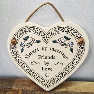 May include: Cream-coloured heart-shaped ceramic plaque with the text "Sisters by marriage Friends by Love". It features a black decorative border and floral details in pink, blue, and green. A brown rope is attached for hanging.