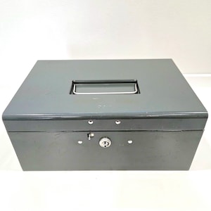 May include: A grey metal cash box with a rectangular form. It has a handle on the top and a key lock on the front. The box is designed for secure storage of money and valuables.