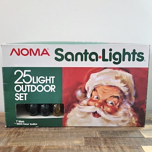 May include: A rectangular cardboard box for a NOMA Santa-Lights 25 light outdoor set. The packaging features a red background with a detailed illustration of Santa Claus and white text. A small cutout window reveals several dark-coloured light bulbs.
