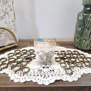 May include: A selection of vintage brass curtain rings, some in a clear plastic bag, and others arranged on a white lace doily. The rings are of various shapes, including round and square. The packaging reads "Drapery Rings with Eye".