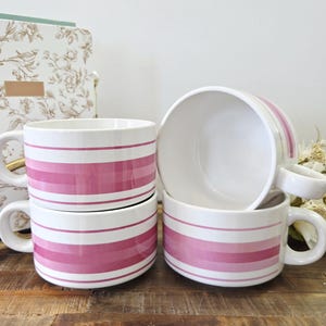 Peut inclure: Quatre tasses en céramique blanche avec des rayures horizontales roses. Les tasses ont une forme arrondie avec une anse sur le côté. Les tasses sont empilées sur une surface en bois.