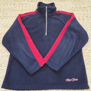 May include: Navy blue fleece pullover with a red stripe design on the sleeves. Features a quarter-zip closure and the text "Race Face" in red script on the lower right. A casual, comfortable top.