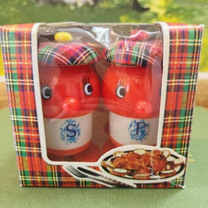 Könnte beinhalten: Ein Set mit Salz- und Pfefferstreuern im Vintage-Stil. Die roten Streuer haben weiße Sockel mit blauen Buchstaben und sind mit Tartan-Kappen versehen. Die Box ist rot, grün und gelb kariert.