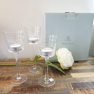 May include: Three clear glass candle holders with etched horizontal lines, each featuring a stem and a circular base. The holders are positioned in front of a light blue PartyLite box and a single white artificial flower with green stems.