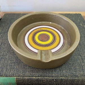 May include: An olive-green ashtray with a central circular design. The design includes concentric rings of brown, white, and yellow. The ashtray has a small notch for cigarettes.
