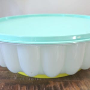May include: A vintage, white plastic mold with a scalloped design, topped with a light teal lid. The base is yellow. This kitchenware item is likely used for making gelatin desserts or molded salads.