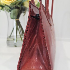 May include: A reddish-brown leather handbag with a stitched design. The bag has two handles and a zipper closure. The bag is next to a vase of yellow roses.
