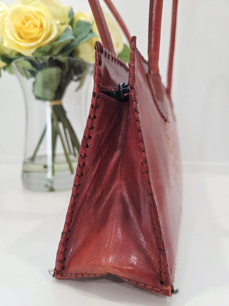 May include: A reddish-brown leather handbag with a stitched design. The bag has two handles and a zipper closure. The bag is next to a vase of yellow roses.