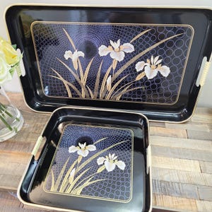 May include: A set of two rectangular black serving trays featuring a central graphic of white iris flowers with long green stems against a dark blue background with a repeating circular pattern. Each tray has gold trim and integrated side handles.