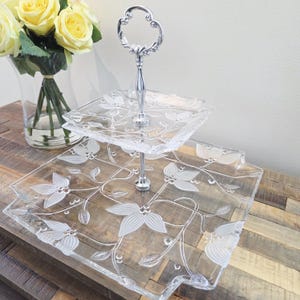 Peut inclure: Un présentoir à gâteaux en verre transparent à deux niveaux avec une poignée argentée. Les plateaux carrés présentent un motif floral. Des roses jaunes dans un vase en verre sont en arrière-plan.