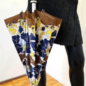 Pu&ograve; includere: Un ombrello vintage con motivo floreale. L'ombrello presenta un bordo marrone, uno sfondo bianco e un motivo di fiori blu e gialli. L'impugnatura &egrave; argentata e nera.