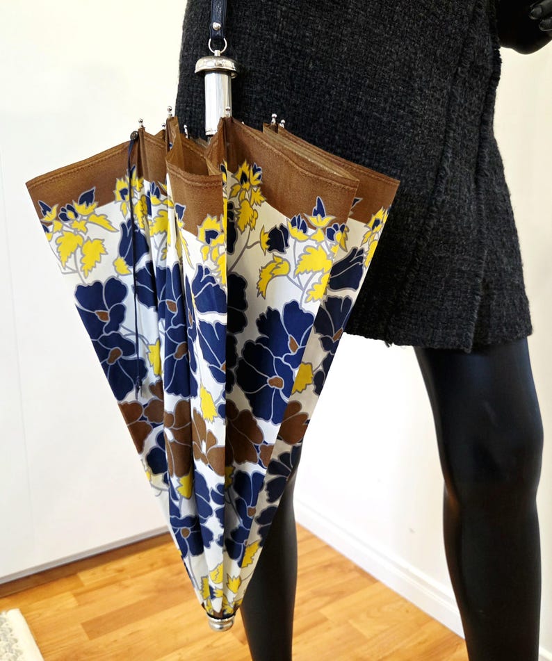 Pu&ograve; includere: Un ombrello vintage con motivo floreale. L'ombrello presenta un bordo marrone, uno sfondo bianco e un motivo di fiori blu e gialli. L'impugnatura &egrave; argentata e nera.