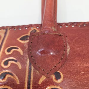 May include: Close-up of a brown leather handbag with a handle. The bag features decorative embossed patterns and stitched detailing. The handle is attached with a leather patch, and the bag appears to be well-worn.