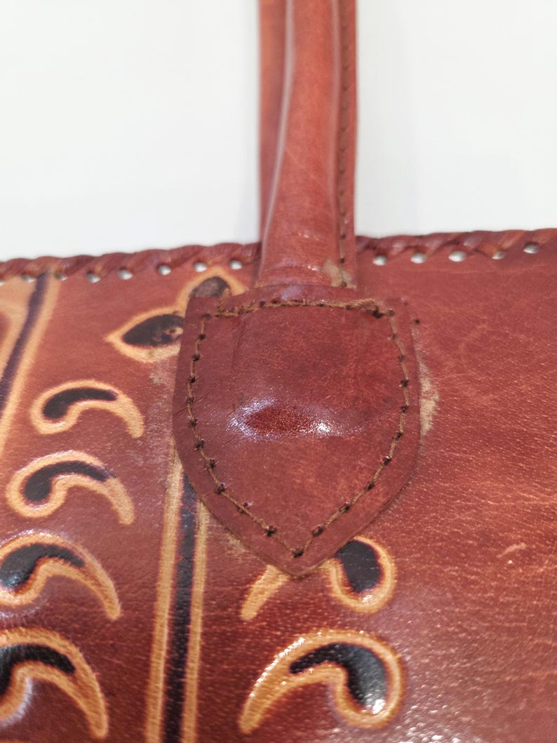 May include: Close-up of a brown leather handbag with a handle. The bag features decorative embossed patterns and stitched detailing. The handle is attached with a leather patch, and the bag appears to be well-worn.