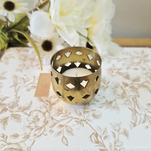 Peut inclure: Un petit bougeoir rond en métal doré avec une finition vieillie et un motif répétitif de découpes en forme de losange. Le bougeoir possède une ouverture sur le dessus et contient une bougie chauffe-plat blanche posée sur une surface à motif floral.