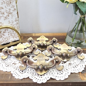 以下が含まれることがあります： 装飾的なデザインの、真鍮製のアンティーク引き出しプル5個。白いレースのドイリーの上に配置されています。引き出しプルは、湾曲したハンドルと装飾的なバックプレートを備えています。ヴィンテージのホームデコレーションアイテムです。