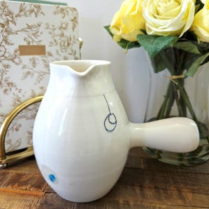 Peut inclure: Une cruche en céramique blanche avec un corps arrondi et une longue poignée. La cruche a un bec verseur et est décorée d'un motif bleu. Un bouquet de roses jaunes est à proximité dans un vase en verre.