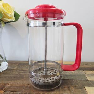 May include: A clear glass French press coffee maker with a red lid and handle. The press has a stainless steel filter and a silver band around the top and bottom. The coffee maker is on a wooden surface.