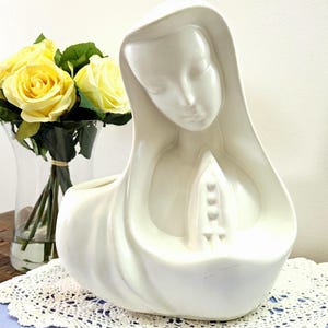May include: A white ceramic statue of a woman with a draped head covering and clasped hands. The statue has a smooth, glossy finish. Yellow roses in a glass vase are in the background.