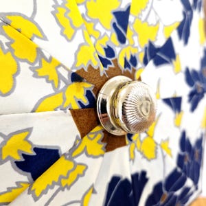 Pu&ograve; includere: Primo piano di un ombrello a fantasia floreale. L'ombrello presenta uno sfondo bianco con disegni floreali gialli, blu e grigi. L'impugnatura &egrave; argentata e gli accenti sono marroni.