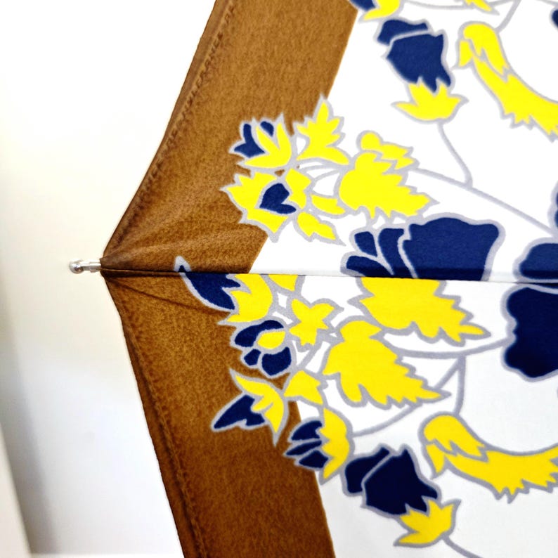 Pu&ograve; includere: Primo piano di un ombrello aperto. L'ombrello ha un bordo marrone e un interno bianco con motivi floreali gialli e blu navy. Il telaio &egrave; visibile nella parte superiore.