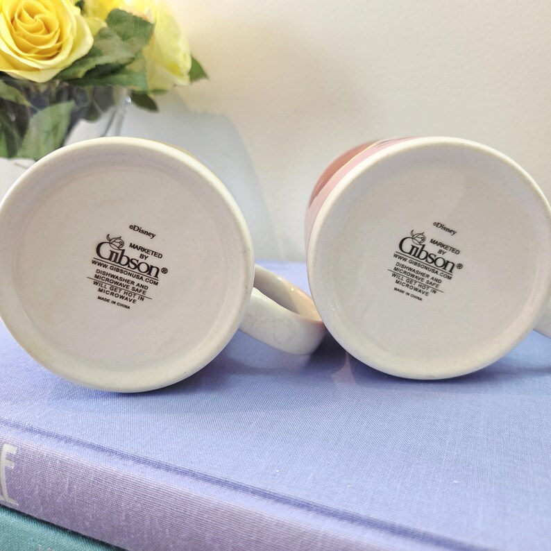 Op de afbeelding: Twee witte keramische mokken met de tekst "Disney" en "MARKETED BY Gibson&reg;" op de bodem. De mokken staan op een lichtpaars oppervlak. De mokken zijn wit met een roze rand.