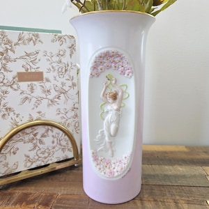 Peut inclure: Un vase en céramique blanc et lavande avec un bord doré. Le vase présente un relief d'une femme dansant sous une arche florale. Les fleurs sont roses et la femme porte une robe blanche. Le vase est sur une surface en bois.