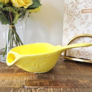 May include: A vibrant yellow ceramic gravy boat with a long handle and a pouring spout. The boat has an embossed floral pattern. It rests on a wooden surface, with a vase of yellow roses in the background. The boat's design is reminiscent of vintage kitchenware.