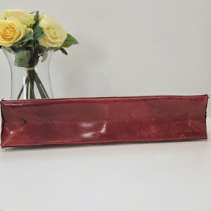 May include: A rectangular, deep red leather clutch with a smooth, slightly glossy finish and visible stitching along the edges. The item is positioned horizontally, showcasing its elongated, structured form and rich, dark crimson color.
