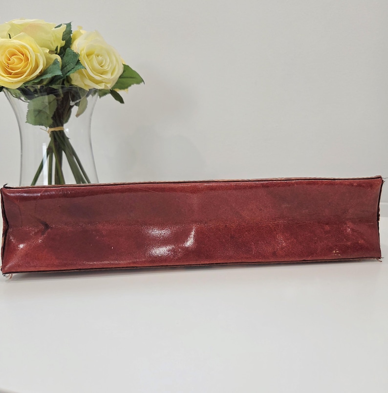 May include: A rectangular, deep red leather clutch with a smooth, slightly glossy finish and visible stitching along the edges. The item is positioned horizontally, showcasing its elongated, structured form and rich, dark crimson color.
