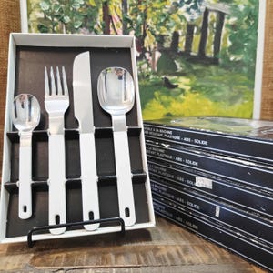 May include: A boxed set of stainless steel flatware with white handles. The set includes a spoon, fork, knife, and teaspoon. The box is black and white, and the background includes a painting and additional boxes.
