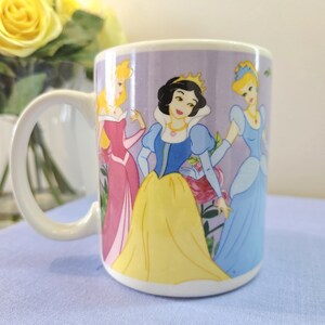 Op de afbeelding: Witte keramische mok met illustraties van Disney-prinsessen Aurora, Sneeuwwitje en Assepoester. De prinsessen zijn afgebeeld in hun iconische jurken tegen een lavendelkleurige achtergrond. De mok heeft een gebogen handvat en een glanzende afwerking.