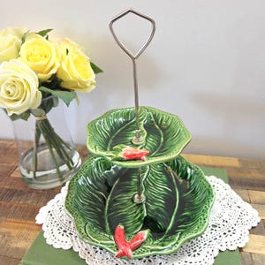 Peut inclure: Plat à deux niveaux avec des assiettes en céramique vertes en forme de feuilles. Les assiettes ont un motif texturé et sont agrémentées de détails de piments rouges. Une poignée en métal argenté s'étend vers le haut.