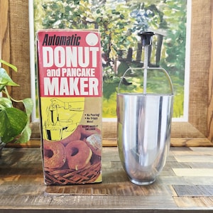 Puede incluir: Caja vintage "Automatic Donut and Pancake Maker" con un dispensador de acero inoxidable. La caja es roja, amarilla y marrón, con texto e ilustraciones. El dispensador tiene un mango negro y un mecanismo de resorte. Se incluye un folleto de recetas.