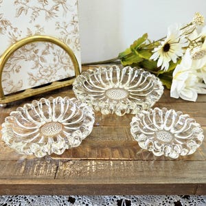 Peut inclure: Trois cendriers en verre transparent en forme de tournesol. Les cendriers sont disposés sur une surface en bois. Un support en métal doré avec un motif floral et un bouquet de fleurs blanches sont en arrière-plan.