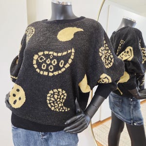 May include: A dark grey sweater featuring gold paisley patterns. The sweater has a round neck and long sleeves. The mannequin is wearing a denim skirt. The design includes various paisley shapes and circular patterns.