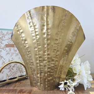 Peut inclure: Grande pièce décorative en laiton antique en forme de coquillage. Le laiton a une surface texturée avec un effet martelé. La pièce est exposée sur une surface en bois avec des fleurs blanches.