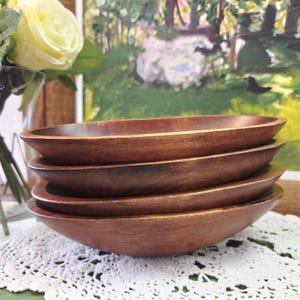 Peut inclure: Une pile de quatre bols ovales en bois. Les bols sont de couleur marron chaud avec un grain de bois visible. Ils sont empilés les uns sur les autres, mettant en valeur leur profondeur et leur forme. Les bols sont sur une nappe en dentelle blanche.