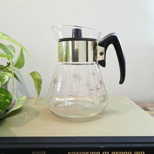 May include: A clear glass coffee carafe with a silver band and black handle. The carafe features a starburst pattern and a black lid. The carafe is on a book.