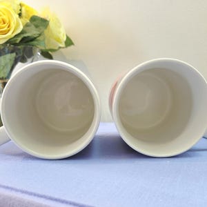 Op de afbeelding: Twee witte keramische mokken met handvatten, van bovenaf gezien, staan ​​naast elkaar op een lichtblauwe ondergrond. De binnenkant van de mokken is wit. Gele rozen in een glazen vaas zijn op de achtergrond te zien.
