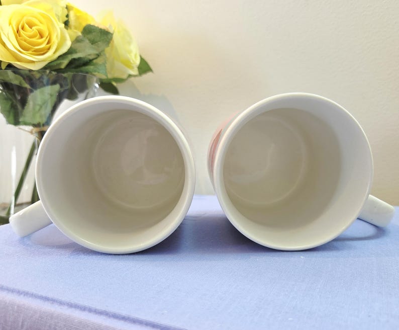 Op de afbeelding: Twee witte keramische mokken met handvatten, van bovenaf gezien, staan ​​naast elkaar op een lichtblauwe ondergrond. De binnenkant van de mokken is wit. Gele rozen in een glazen vaas zijn op de achtergrond te zien.
