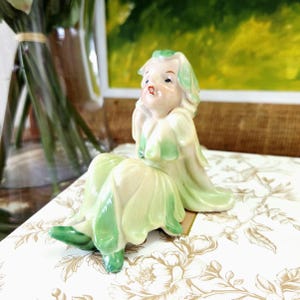 Peut inclure: Figurine en céramique d'une femme assise, vêtue d'une robe et de cheveux verts et crème. La figurine a une expression sereine et repose sur une surface à motifs floraux. C'est un objet décoratif.