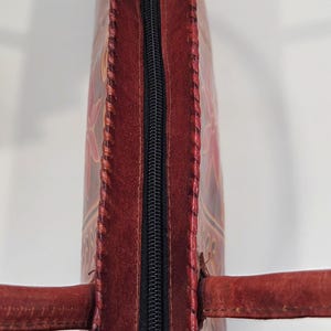 May include: A handcrafted, red leather tote bag with a black zipper closure. The bag features intricate floral designs and stitched edges. The handles are also made of red leather. The bag is a stylish accessory.