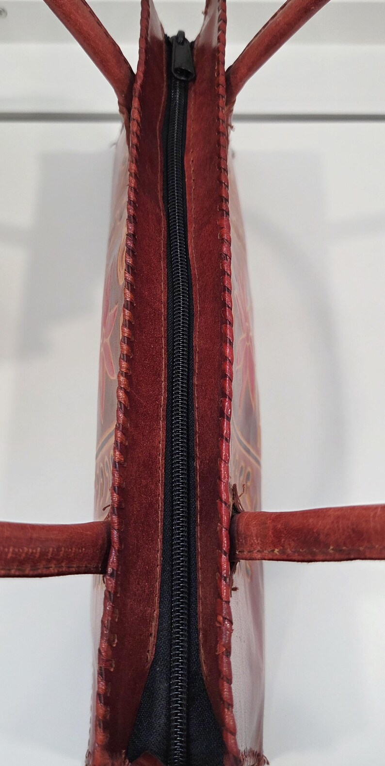 May include: A handcrafted, red leather tote bag with a black zipper closure. The bag features intricate floral designs and stitched edges. The handles are also made of red leather. The bag is a stylish accessory.