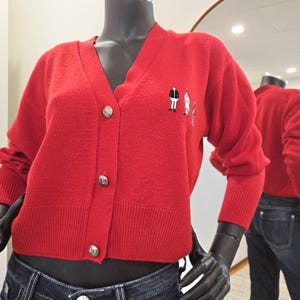 May include: A bright red cardigan sweater with a V-neck and three silver buttons. The sweater has small embroidered figures on the left side. The sweater is displayed on a mannequin.