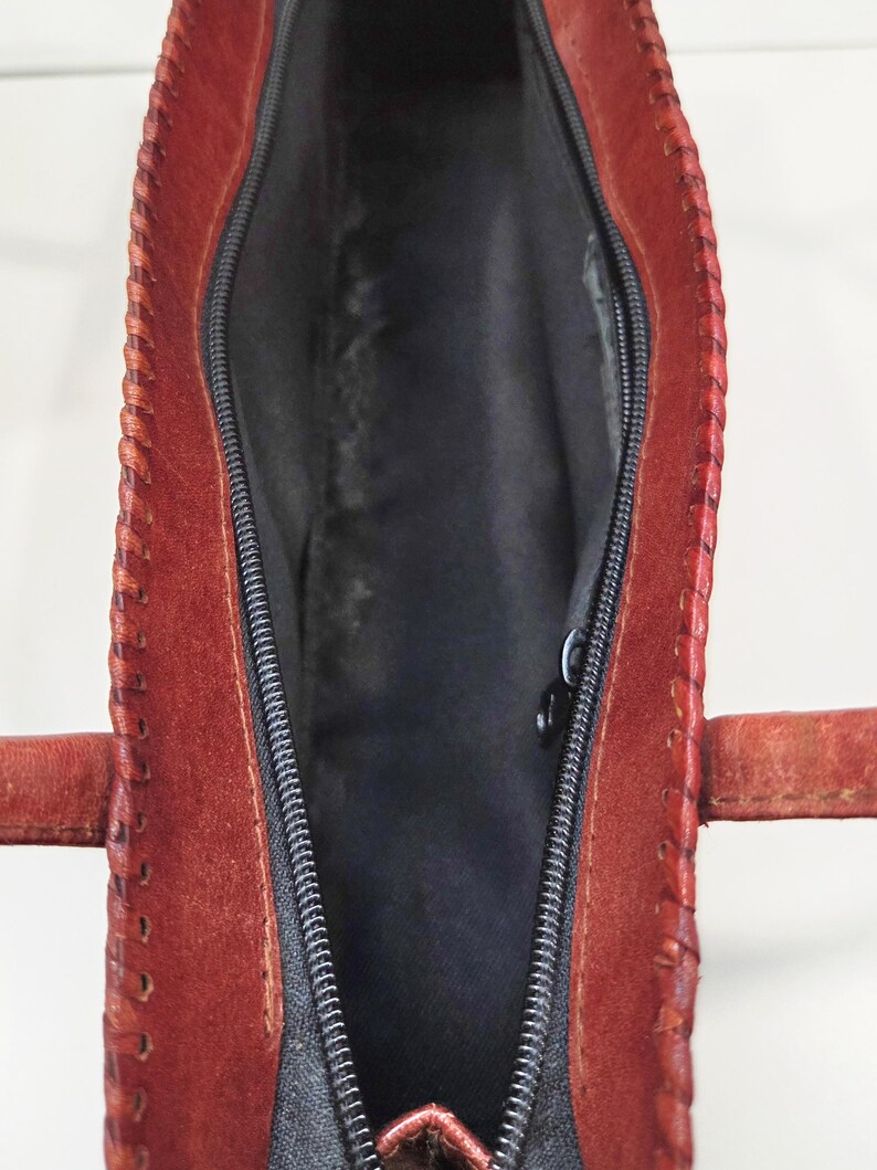 May include: Open view of a red leather handbag with a black zipper and interior. The bag has a stitched design along the edges and two small handles. The interior is lined with black fabric.