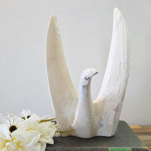 Op de afbeelding: Een witte, abstracte vogelsculptuur met uitgestrekte vleugels. De sculptuur heeft een textuur en staat op een donkergroen boek. Witte bloemen zijn links van de sculptuur gerangschikt, wat een serene, artistieke presentatie creëert.