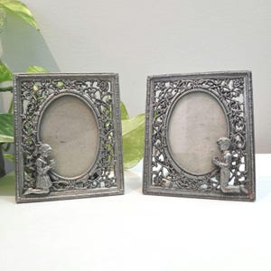 May include: Two silver-toned metal picture frames. Each frame has an oval opening and is decorated with a floral design and a kneeling figure. The frames are rectangular with ornate detailing.