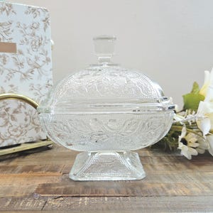 Peut inclure: Un bonbonnier en verre pressé transparent avec un couvercle assorti, présentant un motif floral et végétal en relief sur tout le corps et le couvercle. Le récipient repose sur une base carrée et possède un petit bouton de préhension.